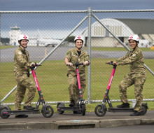Three aviators in camouflage uniform each stand with one food on a pink Flamingo electric scooter. They stand in front of a fence on the outskirts of Base Ohakea with a plane, tarmac and hangers in the background.