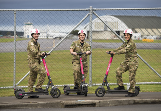 Three aviators in camouflage uniform each stand with one food on a pink Flamingo electric scooter. They stand in front of a fence on the outskirts of Base Ohakea with a plane, tarmac and hangers in the background.