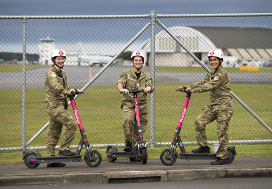 Three aviators in camouflage uniform each stand with one food on a pink Flamingo electric scooter. They stand in front of a fence on the outskirts of Base Ohakea with a plane, tarmac and hangers in the background.