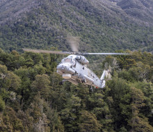 A grey Seasprite helicopter flies in front of forest covered hills, the rotors are blurred slightly and the landing gear is down.