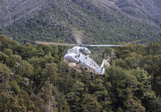 A grey Seasprite helicopter flies in front of forest covered hills, the rotors are blurred slightly and the landing gear is down.