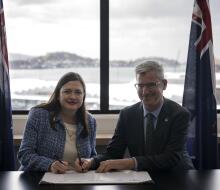 Two people co-sign a document between the Australian and New Zealand flags with a large bright window behind them.