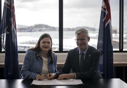 Two people co-sign a document between the Australian and New Zealand flags with a large bright window behind them.