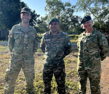 Three soldiers in stand side-by-side in uniform, looking at the camera and smiling. It is a sunny day and blue sky. They are standing on grass and there are trees in the background. 