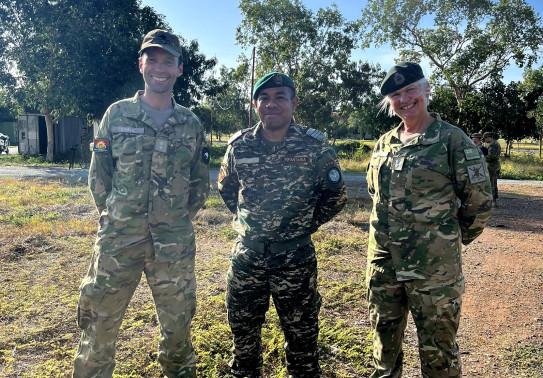 Three soldiers in stand side-by-side in uniform, looking at the camera and smiling. It is a sunny day and blue sky. They are standing on grass and there are trees in the background. 