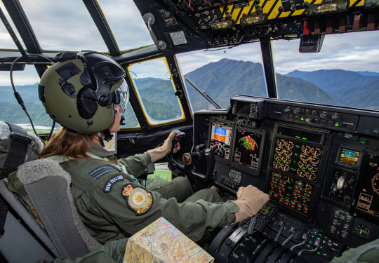 A female pilot wearing a green suit and helmet inside the flight deck of a Hercules aircraft. 