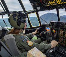 A female pilot wearing a green suit and helmet inside the flight deck of a Hercules aircraft. 