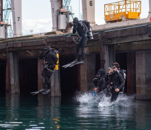 Four divers jump into the water from the wharf at the Port of Tauranga. They are all wearing full dive apparatus, including wet suits, tanks and fins.