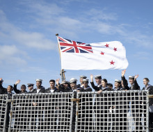 Sailors wave from behind a netted fence on a ship underneath the Navy flag as it blows in the wind. The flag has a Union Jack and four stars on it