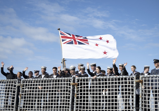 Sailors wave from behind a netted fence on a ship underneath the Navy flag as it blows in the wind. The flag has a Union Jack and four stars on it