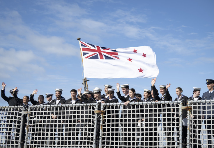 Sailors wave from behind a netted fence on a ship underneath the Navy flag as it blows in the wind. The flag has a Union Jack and four stars on it