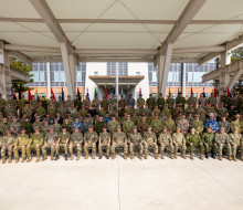 Personnel from a variety of militaries sit for a large group photo in a covered outdoor area. All personnel are wearing the uniform of their military and most of these are a camouflage green.