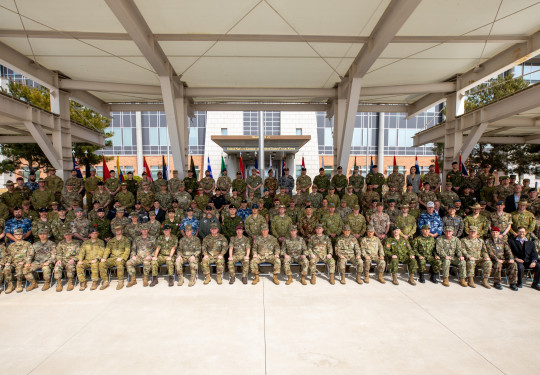 Personnel from a variety of militaries sit for a large group photo in a covered outdoor area. All personnel are wearing the uniform of their military and most of these are a camouflage green.
