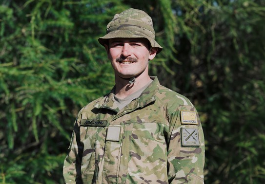 A young male soldier with a moustache, smiles and looks at the camera. He is wearing a camouflaged jungle hat tied under his chin and a camouflaged uniform. He stands in front of a leafy pine tree that fills the frame behind.