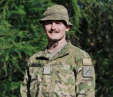 A young male soldier with a moustache, smiles and looks at the camera. He is wearing a camouflaged jungle hat tied under his chin and a camouflaged uniform. He stands in front of a leafy pine tree that fills the frame behind.