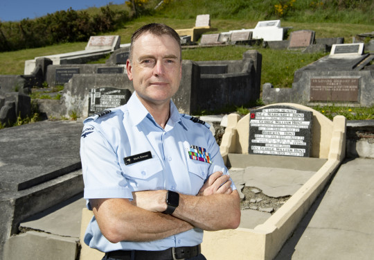 An Airmen with short brown hair, wearing a light blue Air Force shirt and with arms crossed stands in front of a grave at a cemetery. 
