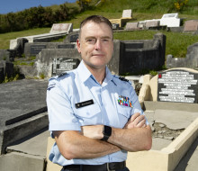 An Airmen with short brown hair, wearing a light blue Air Force shirt and with arms crossed stands in front of a grave at a cemetery. 