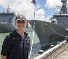 A sailor stands smiling on a sunny day, with some clouds. There are two ships in the background. 