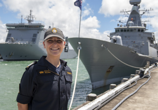 A sailor stands smiling on a sunny day, with some clouds. There are two ships in the background. 