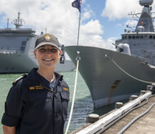 A sailor stands smiling on a sunny day, with some clouds. There are two ships in the background.