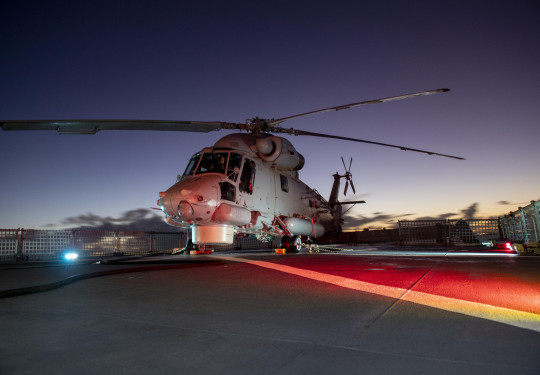 Seasprite helicopter on the deck of HMNZS Otago lit by red lights in the early morning light.
