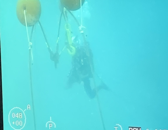 A screen showing the ROV's perspective of a diver as they inspect the plane and secure the plan with strops.