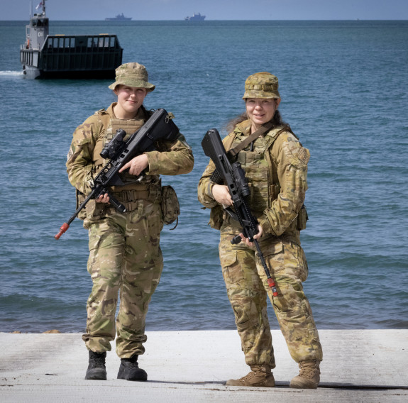 Two soldiers stand side-by-side in uniform holding weapons. In the background is water and a military craft. 