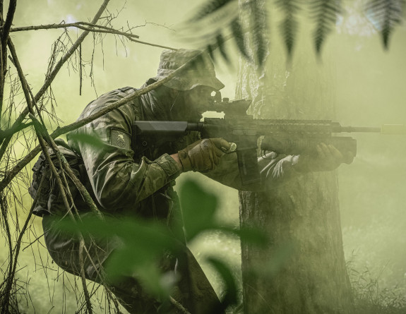 A soldier crouches in the bush, carrying a weapon like they are moving from left to right. The image is very green looking. 