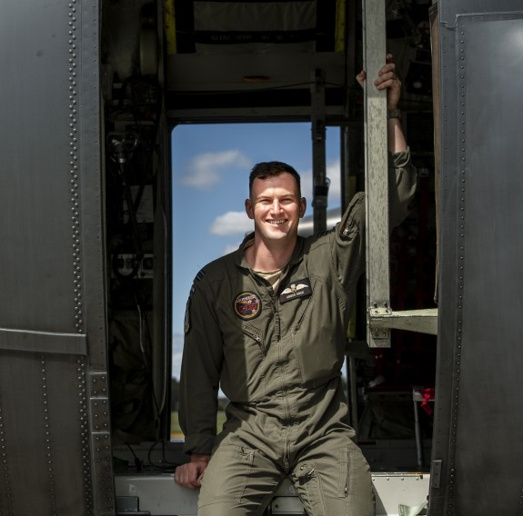 Clean shaven mania a green flight suit sitting in the open side door of a helicopter. The door on the other side is also open and there are blue skies beyond.