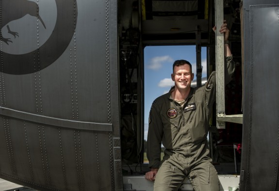 Clean shaven mania a green flight suit sitting in the open side door of a helicopter. The door on the other side is also open and there are blue skies beyond.