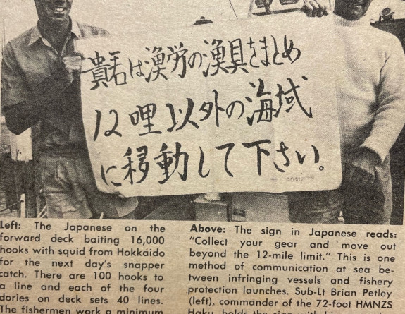A newspaper clipping of showing an image of two navy sailors hold a sign with Japanese writing on it which requests fishermen 'move out beyond the 12-mile limit'.