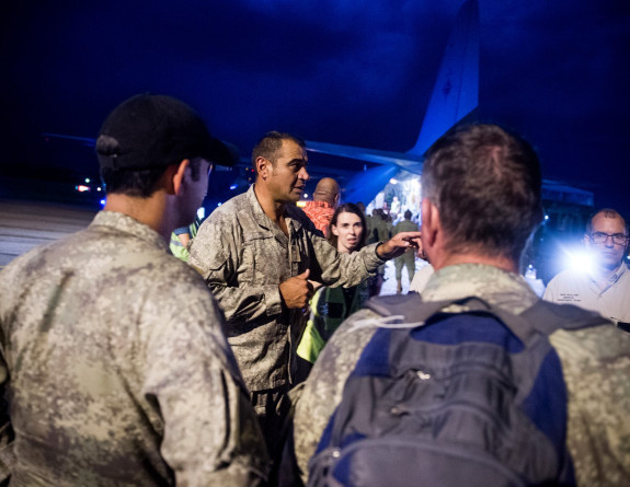 Three New Zealand soldiers in dress fatigues and a man from another government agency are the visible part of a huddle on the tarmac of an airport at night, two soldiers have their back to the camera, they frame te third soldier who is speaking and gestur