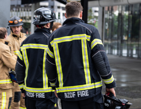 Two NZDF firefighters head to the tent to get ready for their run. They are both wearing black jacket and pants with high visibility and reflective strips. On the back of the jacket reads 'NZDF FIRE'.