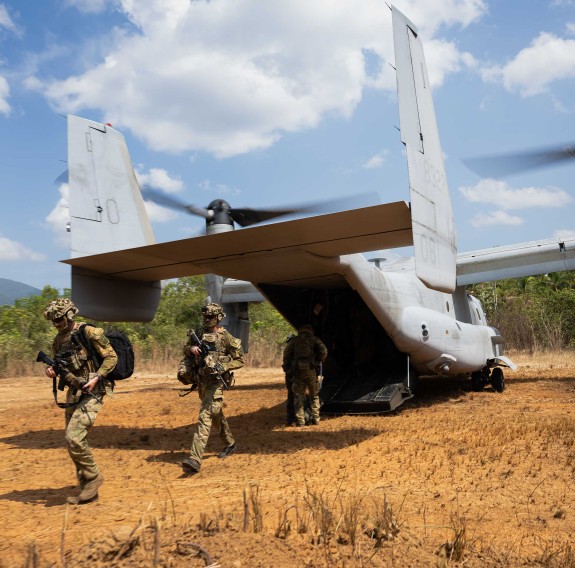 Uniformed soldiers walk off a V-22 Osprey aircraft, the ground is dry, there is blue sky with some clouds. 