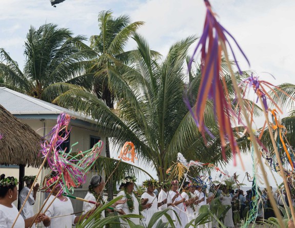 A grey helicopter flies over palm trees and people enjoying festivities, women dressed in white dresses with flags flying.