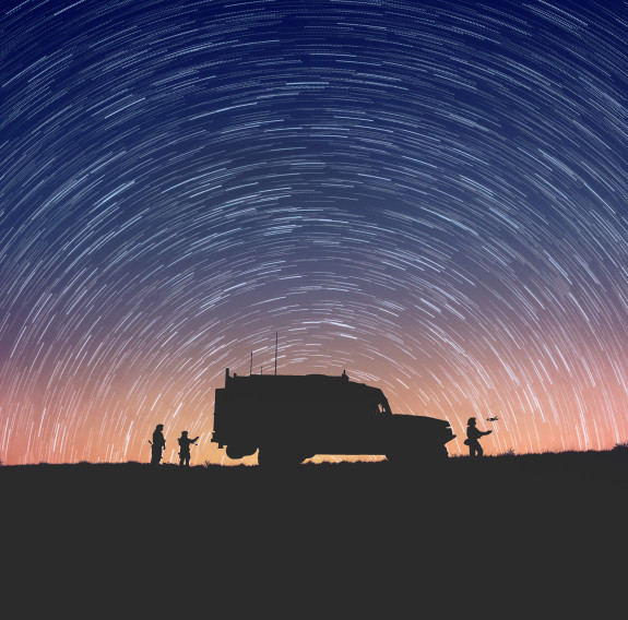 A military vehicle and three personnel stand on the horizon as star trails surround them. The horizon has colours of the sunset, whereas the top of the photo in the stars is a dark blue. 