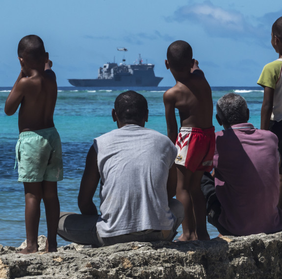 Fijian locals watch from the shore as a Seasprite helicopter lifts an under-slung load from the flight deck of HMNZS Canterbury.