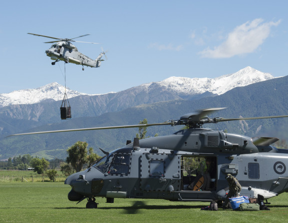 An NH90 helicopter operates from 'LZ High' in Kaikoura, following an Earthquake on 14 Nov 16, while a Seasprite helicopter delivers an underslung load in the background.