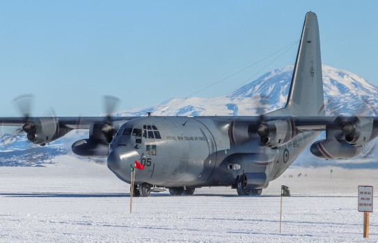 A large grey aircraft with four propellors sits on the ice in Antarctica with large snow covered mountains in the background.