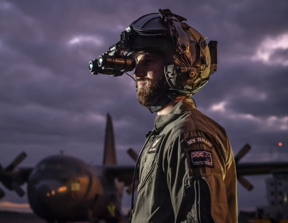 An aircrew member of a C-130H(NZ) Hercules aircraft wearing night vision goggles at dusk in front of the aircraft on the flightline