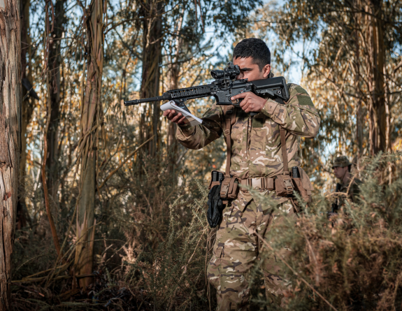 A soldier patrols through dense bush and looks through the scope of the modular assault rifle