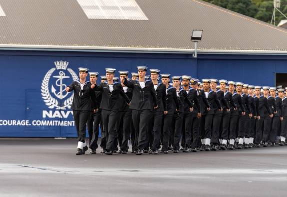 Royal New Zealand Navy recruits march on during a graduation with the Navy logo and values on a wall behind them.  