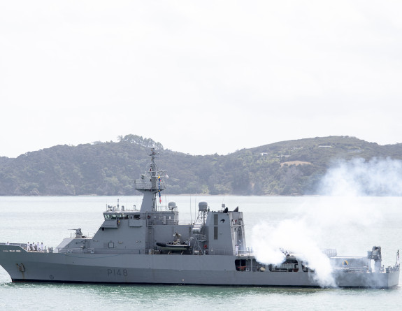 A gun salute is fired on the flight deck of HMNZS Otago on a cloudy day