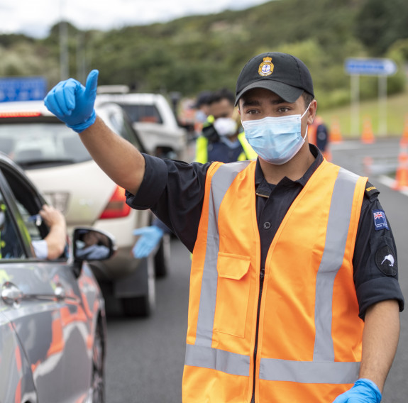 A navy sailor wearing high vis and a mask gives the thumbs up to a vehicle during a checkpoint inspection in Auckland