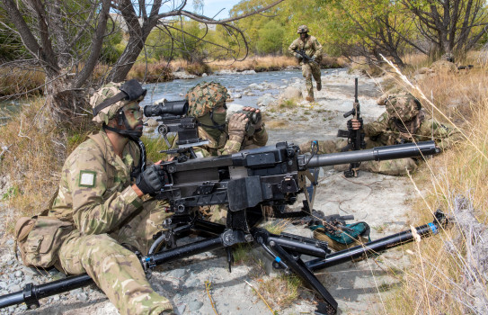 A shooter and spotter sitting together near a riverbed operate a 40mm Grenade Machine Gun