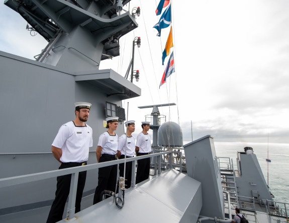 Sailors stand in a row on the ship with flags in the background