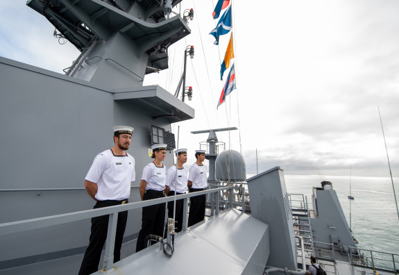 Sailors stand in a row on the ship with flags in the background