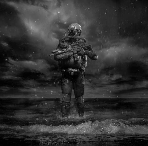 The image is black and white. A soldier stands in a wetsuit and full military gear for the water holding a water. The soldier is holding a weapon.