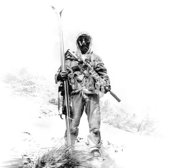 The image is black and white. A soldier stands in front of a snow-capped mountain range, this area is faded white. The soldier is in full uniform and wearing hood up as well as snow goggles. The soldier has a weapon across their chest and is holding skis.