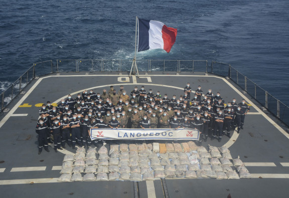 At sea, personnel stand on a ships flight deck, lined up alongside bags of seized narcotics.